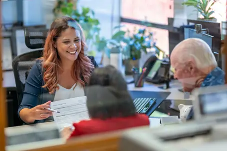 A Hispanic woman sits at her desk at work and smiles while reviewing paperwork with a senior adult couple.