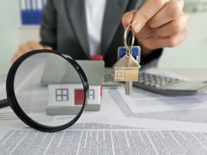 A person holds a set of house keys above a miniature house model while paperwork, a magnifying glass